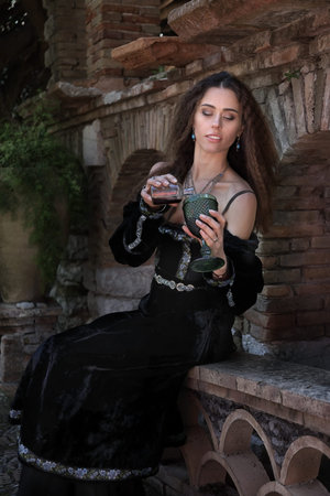 Young beautiful woman in medieval dress pours poison into a glass of wine in the inner courtyard of...