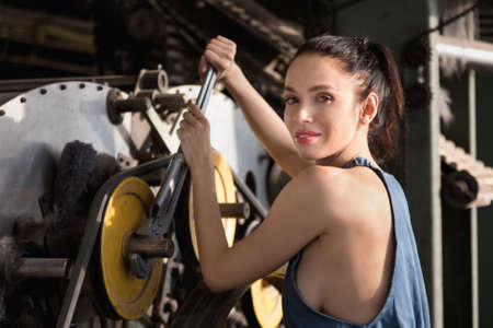 Beautiful brunette girl dressed in overalls repairs a spinning machine with an adjustable wrench in a factoryの写真素材