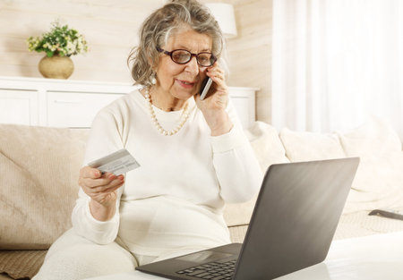 An old woman sitting on a sofa in the living room makes an order using a credit card over the...
