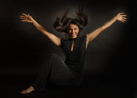 Portrait of a young beautiful woman with outstretched arms sitting in a black evening dress in a...