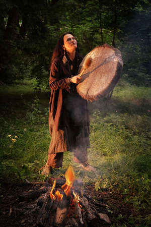 Woman dressed in shaman costume playing on shaman frame drum near large fire in forest at duskの写真素材