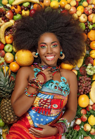 Young beautiful African American Black woman with afro hairstyle smiles at the camera while lying among fruits in the studioの写真素材