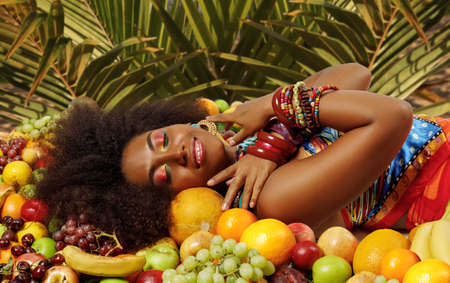 Young lovely African American Black woman smiling with closed eyes while lying among fruits on a background of palm leavesの写真素材