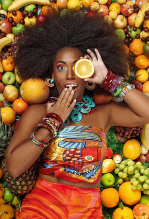 Young beautiful African American Black woman with afro hairstyle closing one eye with a slice of orange while lying among fruits in the studioの写真素材