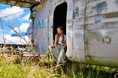 Young nice woman in pin up style clothes stands by an abandoned plane in a field among the grass on a sunny summer dayの写真素材