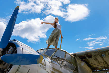 Pretty young adult woman in pin up style clothing stands on an abandoned plane in a field on a sunny summer day against a cloudy skyの写真素材