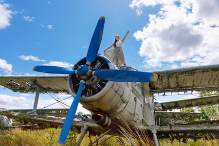Cute young adult woman in pin up style clothing stands on an abandoned plane in a field on a sunny...