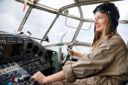 Young beautiful woman in a helmet and pilot suit sitting at the opened door inside a small plane and waving to someone with her handの写真素材