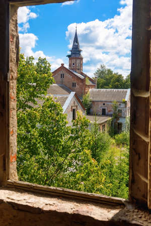 View of the old abandoned Catholic Church overgrown with greenery and trees from the broken window of an abandoned houseの写真素材