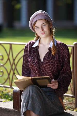 Young pretty student girl in retro clothes is preparing for classes by reading a textbook outdoorsの写真素材