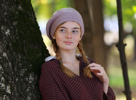 Portrait of a cute freckled girl with a dreamy look in her eyes leaned against a tree in a city park on a sunny summer dayの写真素材