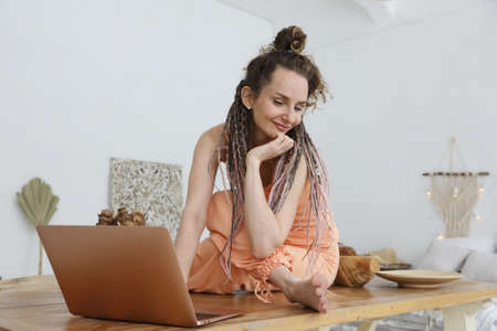 Young woman is working on a laptop sitting on a large wooden table in the living room at homeの写真素材