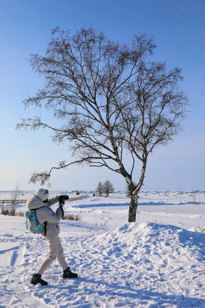 Female photographer takes a picture of a winter landscape on the shore of a frozen lakeの写真素材