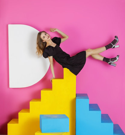 Young beautiful woman in a black dress poses in a studio sitting on colored stairs against a pink wallの写真素材