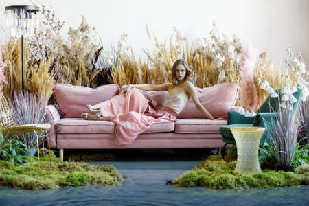 Fantasy concept with a young woman sitting on a sofa in a room in the middle of tall grass with the floor flooded with waterの写真素材