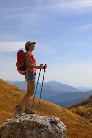 Female tourist with backpack stands on the top of a mountain and admires the beautiful views of the mountains on a sunny autumn dayの写真素材
