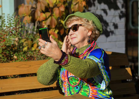 An eighty-year-old grandmother in bright clothes takes a selfie on the phone sitting on a bench outdoors on an autumn sunny dayの写真素材