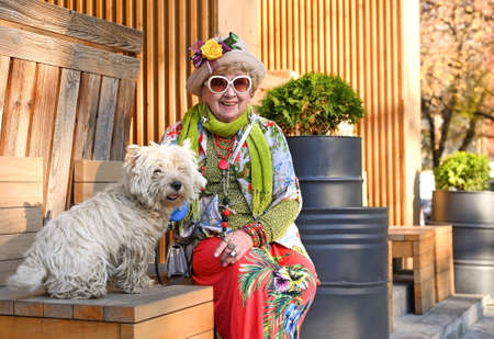 A brightly dressed granny in a hat and sunglasses is sitting on a city bench with her dogの写真素材