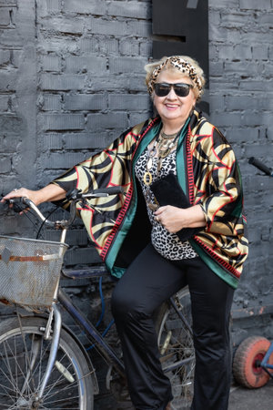 An elderly retired woman in sunglasses and bright clothes poses with a bicycle outdoorsの写真素材