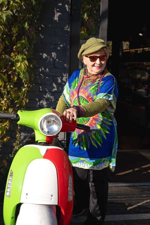 An elderly cheerful woman in bright clothes standing next to the scooter on an autumn sunny dayの写真素材