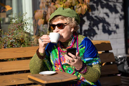 An eighty year old grandmother with a smartphone drinks coffee in a outdoor cafe on a sunny autumn dayの写真素材
