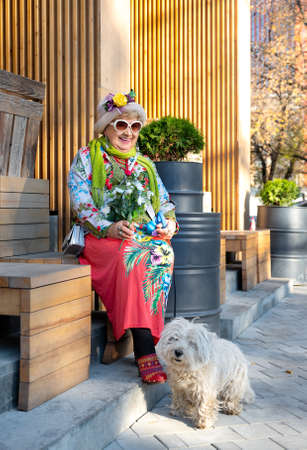 Brightly dressed cheerful eighty year old granny in a hat and sunglasses sitting on a city bench, her dog is sitting next to herの写真素材