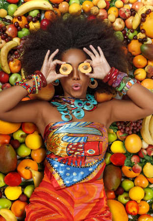 Young beautiful black woman with afro hairstyle closing her eyes with apricots fruit while lying among fruits in the studioの写真素材
