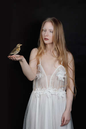 Conceptual art portrait of a young blonde girl with artistic makeup and with a blackbird on her arm on a black backgroundの写真素材