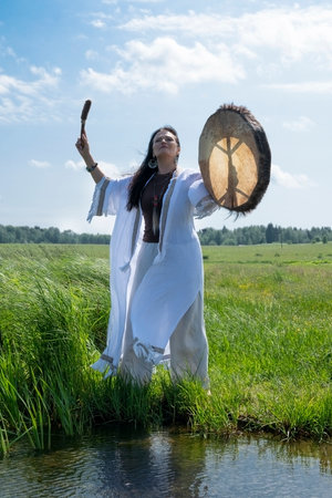 Shaman in the white dress bangs a tambourine on the shore of a lake in natureの写真素材