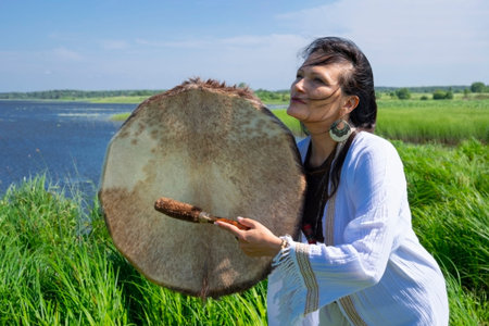Female shaman in the white dress bangs a tambourine on the river bank in nature