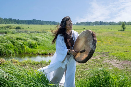 Beautiful female shaman in a trance in a white dress bangs a tambourine on the shore of a lake in...