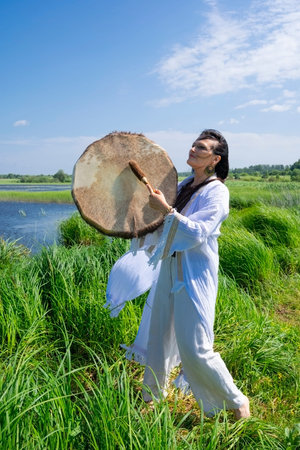 Female shaman in the white dress bangs a tambourine on the river bank in nature
