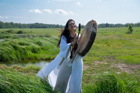 A female shaman in a trance in a white dress bangs a tambourine on the shore of a lake in natureの写真素材