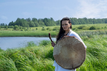 Female shaman in the white dress bangs a tambourine on the river bank in natureの写真素材