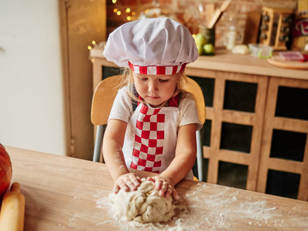 Little girl in cooking chef clothes enjoys kneading the dough. Girls at the kitchen. Family housekeeping.の写真素材