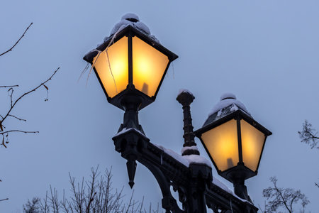 Vintage snow-covered street lamps shine warm light on winter evening.の写真素材