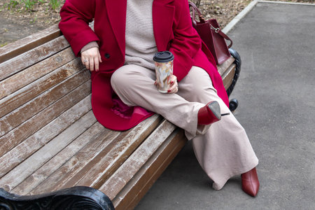 Woman in coat rests on city bench, suffering from discomfort of high heels. Pleasing fashion, wanting to be liked.の写真素材