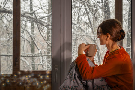 Young woman in cozy plush home-made pajamas sits with cup by window and admires winter landscape with snow-covered trees. Christmas timeの写真素材