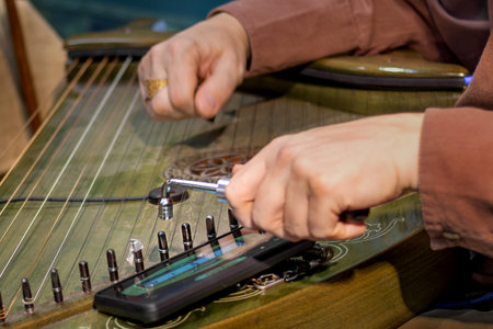 Close-up of mans hands tuning traditional stringed instrument using tuning tool and smartphone app....