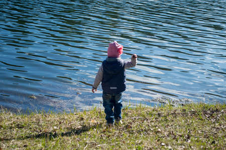 A child (girl) stands on the shore of the lake and throws a lump (stone) into the lakeの写真素材