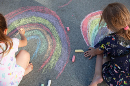 Girls draw a rainbow on the asphalt with colored chalk. Sidewalk. Outdoor kids activities. Drawing...
