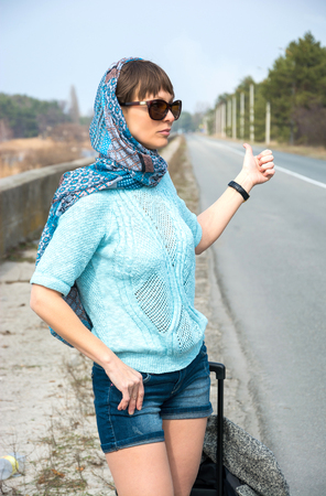 Young woman with a suitcase is hitchhiking on the road pointing her thumb upの写真素材