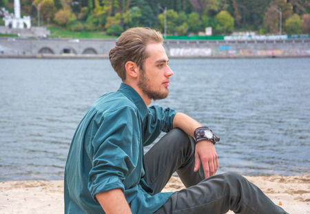 Young man is sitting on the sand near a riverの写真素材
