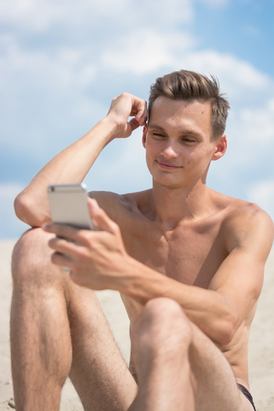 Young athletic man with cellphone on a sky background, on the beachの写真素材