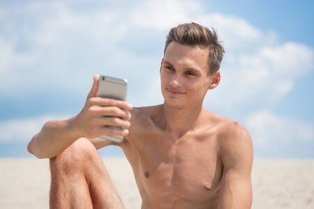 Young athletic tanned man with cellphone on a sky background, on the beach. He is makes selfie and smiling.の写真素材