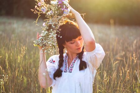 A young woman in a long white embroidered shirt puts on a wreath of wild flowers in a meadow at sunset. Girl in ancient national clothes.の写真素材