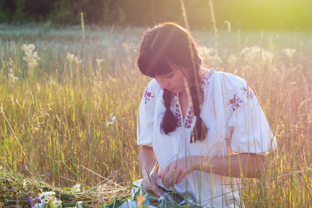A young woman in a long white embroidered shirt weaves a wreath of wild flowers in a meadow at sunset. Girl in ancient national clothes. Light effect from the sun's raysの写真素材