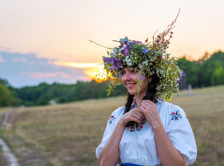 Portrait of a young woman in a long white embroidered shirt and in a wreath of wild flowers stands in the field at sunset. Girl in ukrainian national clothes.の写真素材