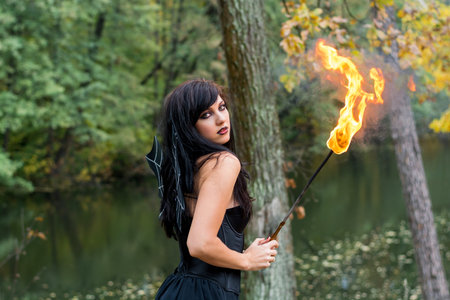 Young brunette women with Halloween witch makeup stands with a torch in a black dress standing in a...