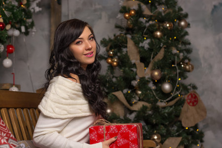 A young woman holds a gift near the Christmas tree. The girl smiles sitting with a gift for Christmasの写真素材